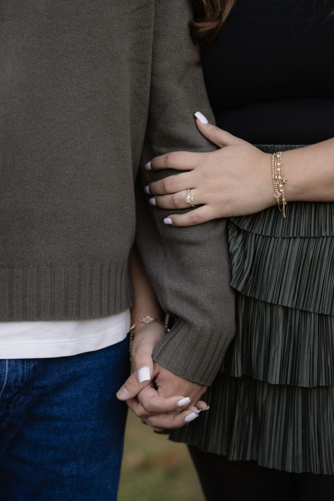a couple holding eachother's hands while standing near eachother. The woman's engagement ring is showing and she is wearing several bracelets. They are posing for their engagement photos and this was a detail shot taken by Amelia and Matt photo