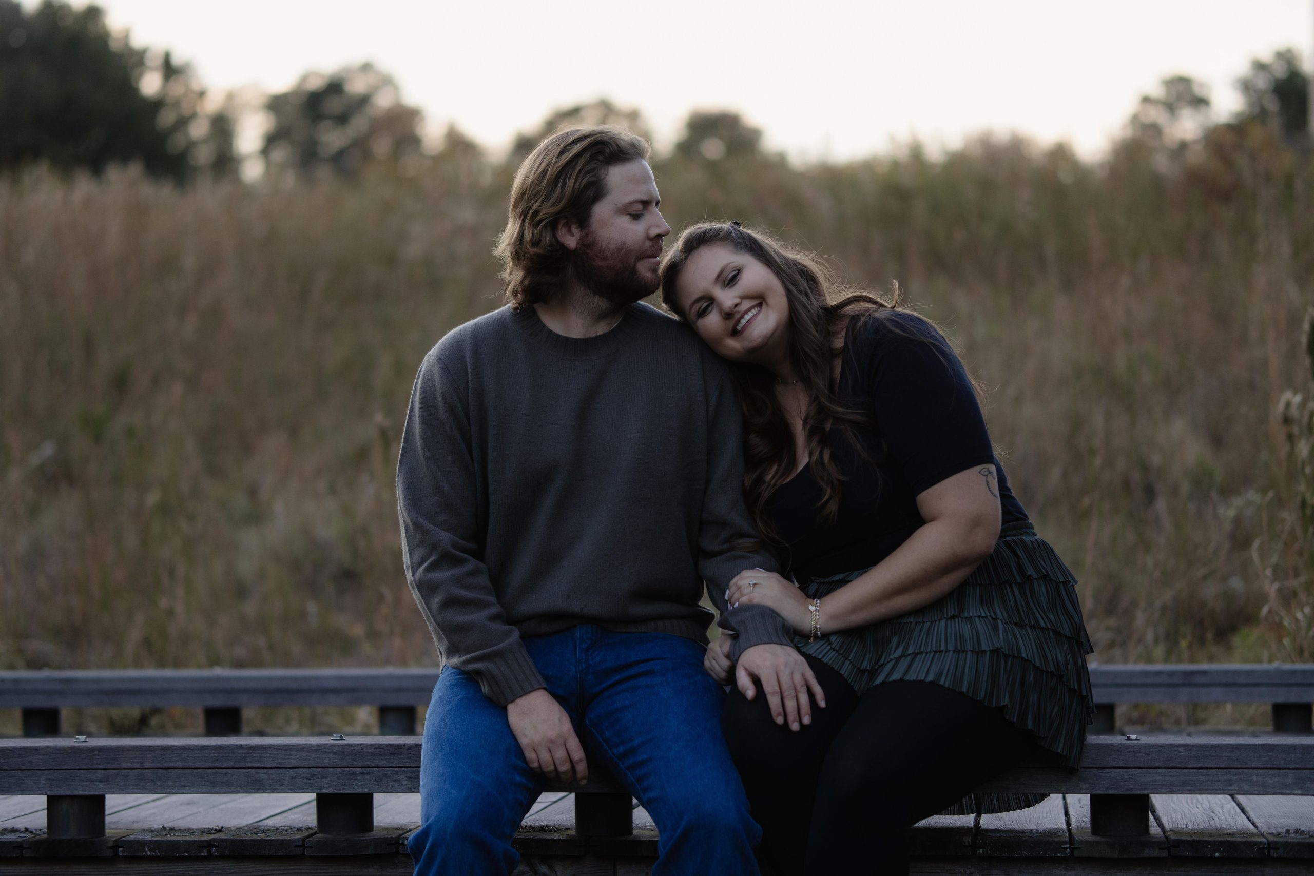 A couple sitting and leaning on eachother in front of some tall grass in their engagement session during golden hour at the NC Art Museum in Raleigh NC