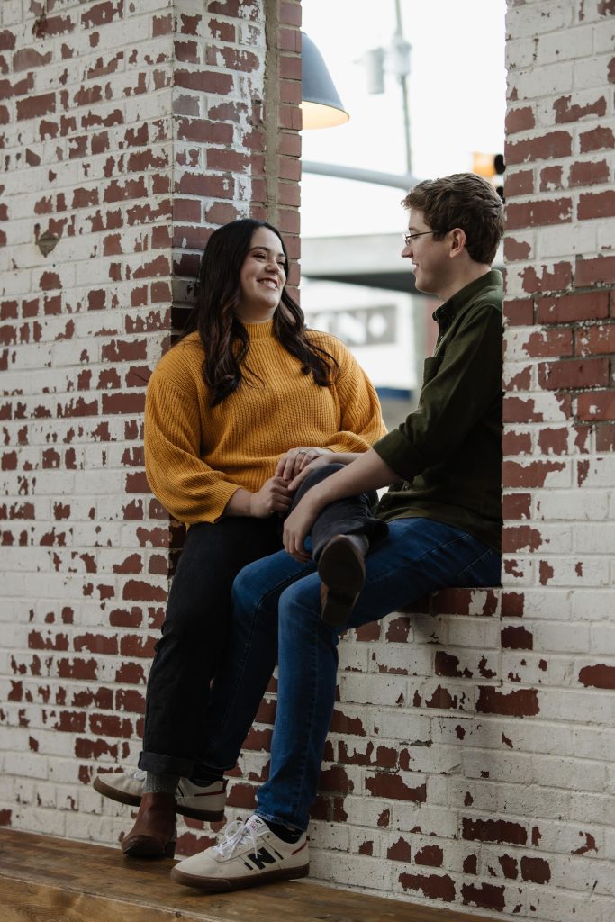 Couple sitting near a brick wall looking sweetly at each other - Downtown Raleigh NC Engagement Session