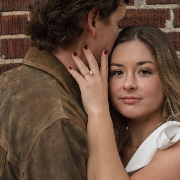 A girl looking into the camera with her new engagement ring showing as she holds her fiancé's face during an engagement session in downtown Raleigh NC
