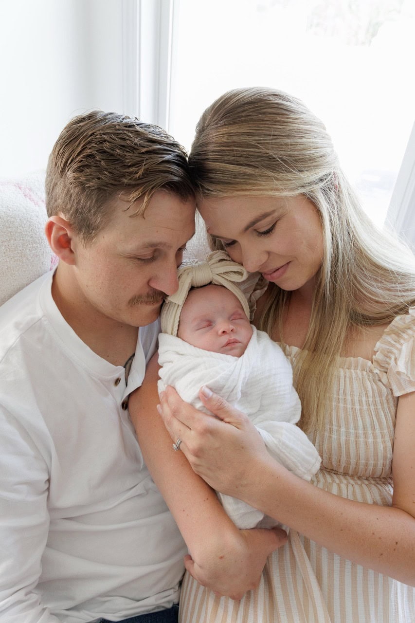 Family sitting on a couch toghether holding their newborn baby infront of a window with sun shining in during their in home lifestyle newborn photography session