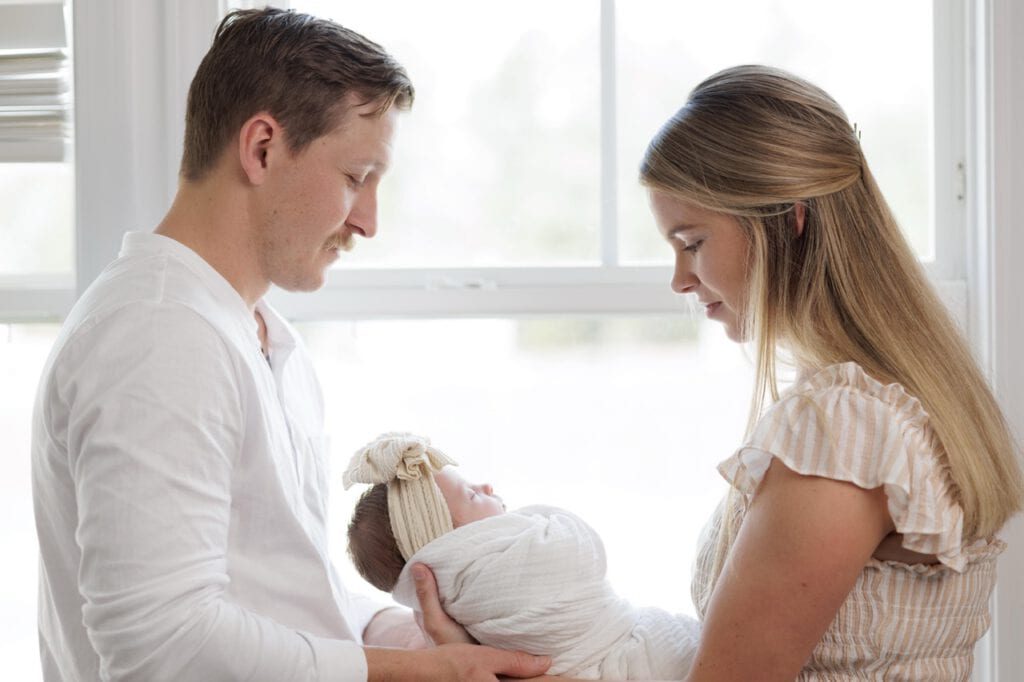 Mom and Dad standing in front of a window looking at their newborn baby in an in-home newborn photography session in Raleigh NC