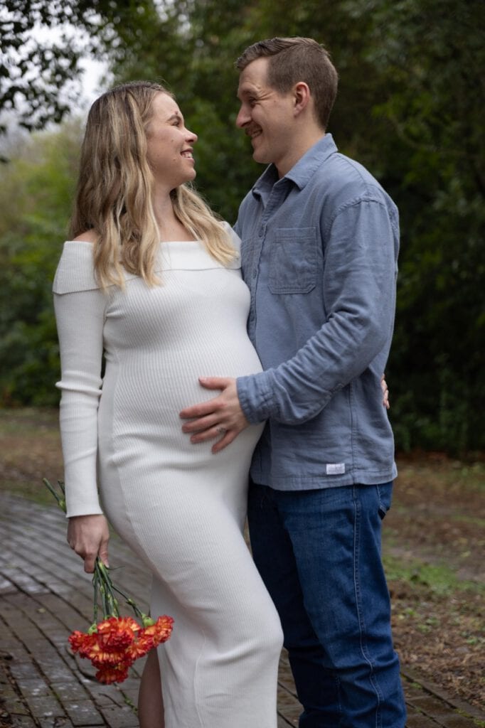 a husband and wife looking sweetly at each other during their Maternity Photoshoot at Oak View Park in Raleigh NC. The husband has his hand of his wife's baby bump and she is holding flowers