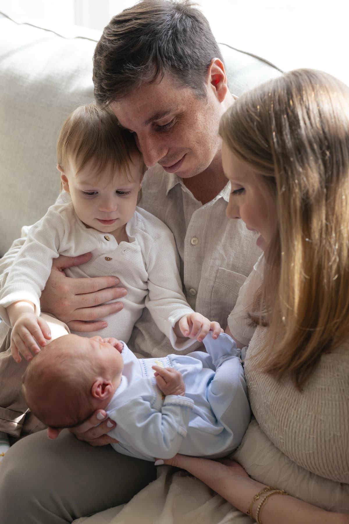 Family sitting on the couch togeher looking down at their newborn baby. Dad is holding his son as mom holds the baby during their in-home lifestyle newborn photography session