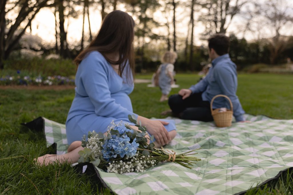 mom and dad sitting on a blanket at WRAL Azalea gardens while their daughter plays in the field