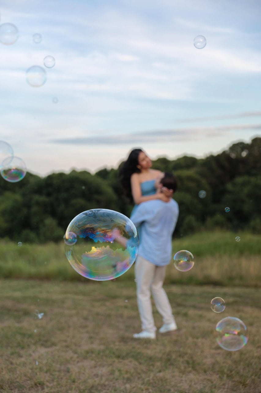 A couple dancing in front of bubbles blown by raleighbubbleman at the ncma in Raleigh. There is tall grass in the background and it is golden hour