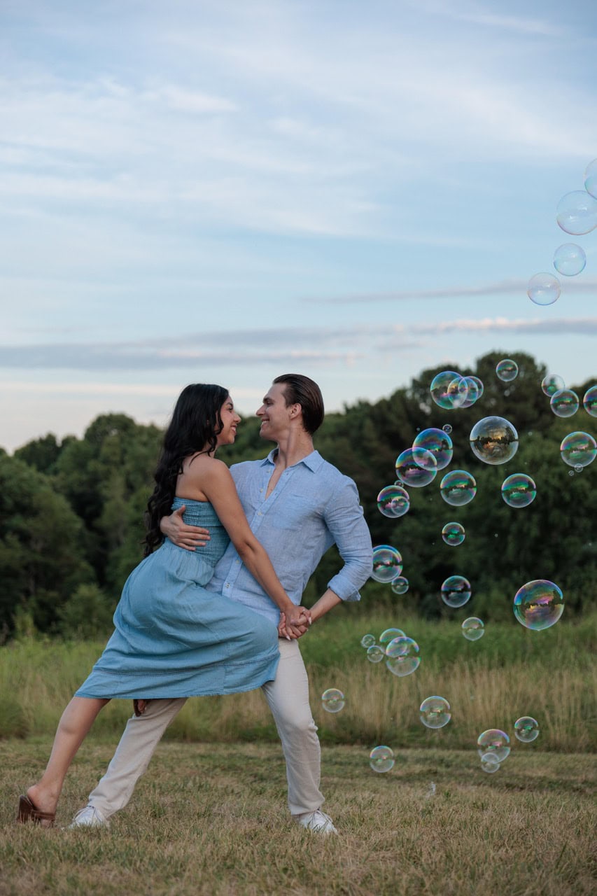 A couple dancing while there are bubbles floating near them in a field of tall grass for their anniversary photoshoot in Raleigh NC