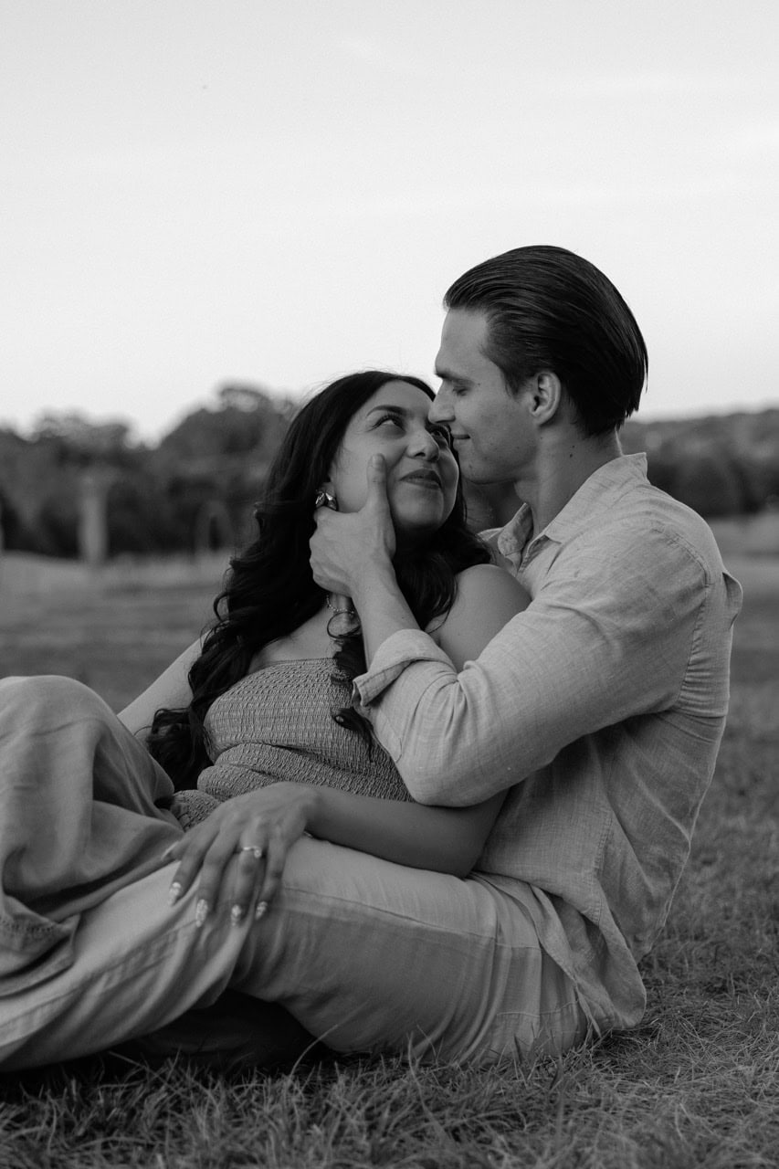 a black and white photo of a couple about to kiss during their anniversary photoshoot for their 1 year. They are sitting is a filed of tall grass