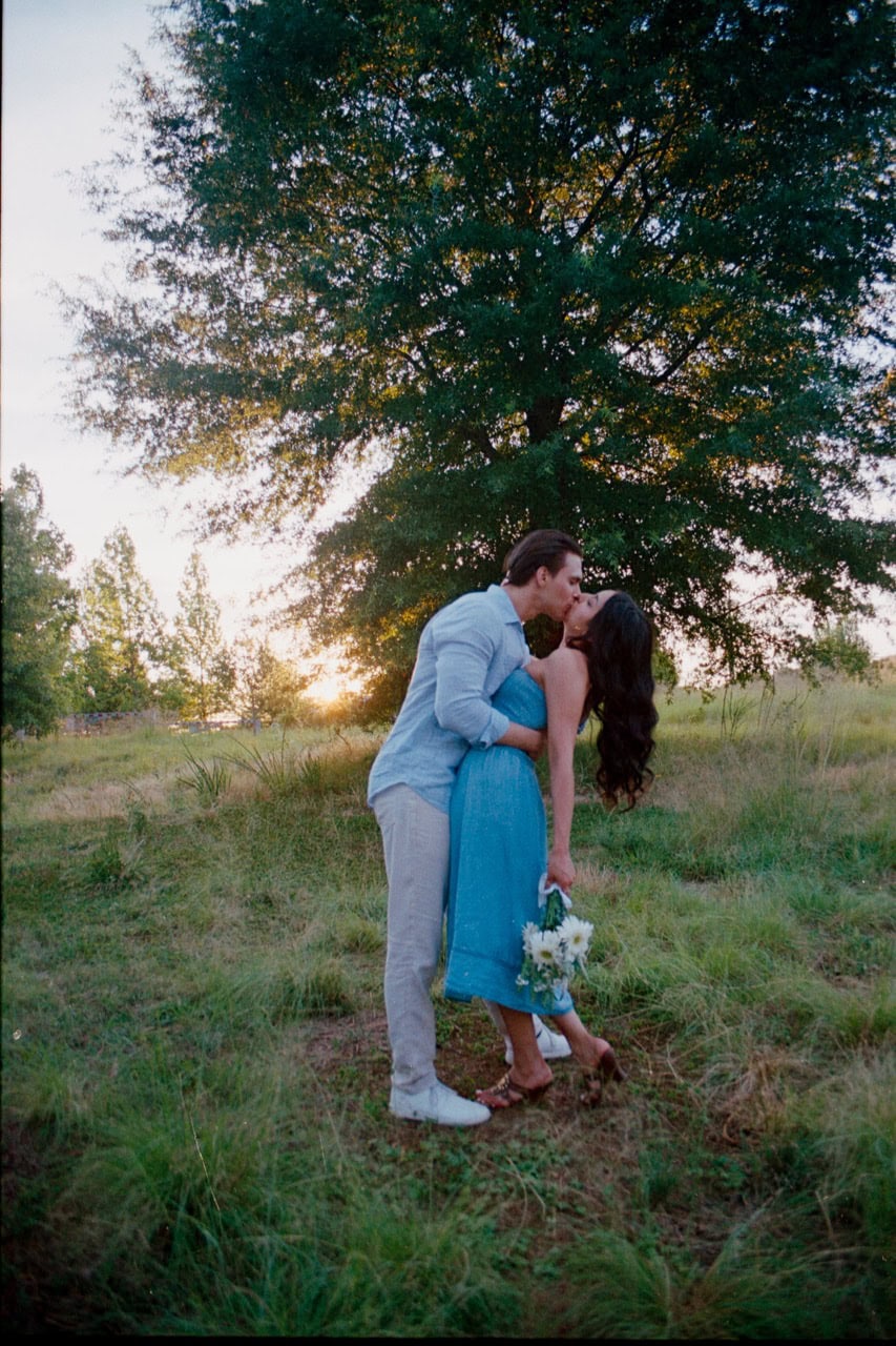 a 35mm film photograph of a couple kissing in front of the sunset in tall grass in front of trees