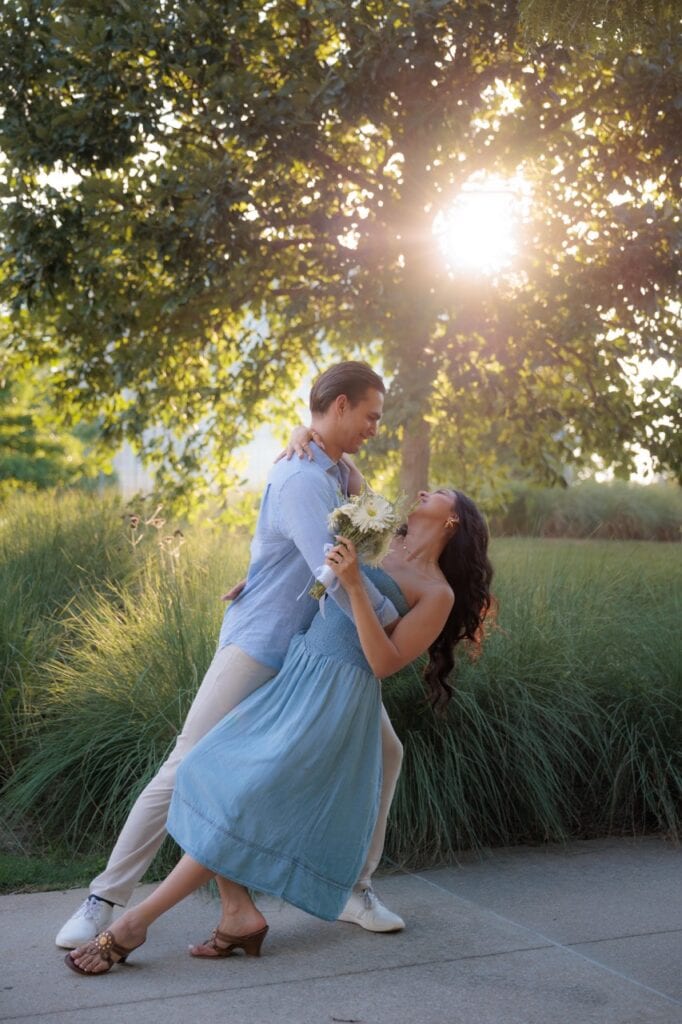 a couple doing a dip during their couples photo shoot with Amelia and Matt Photo. She is holding onto a white bouquet while the golden hour sun is shining through a tree at the NCMA in Raleigh