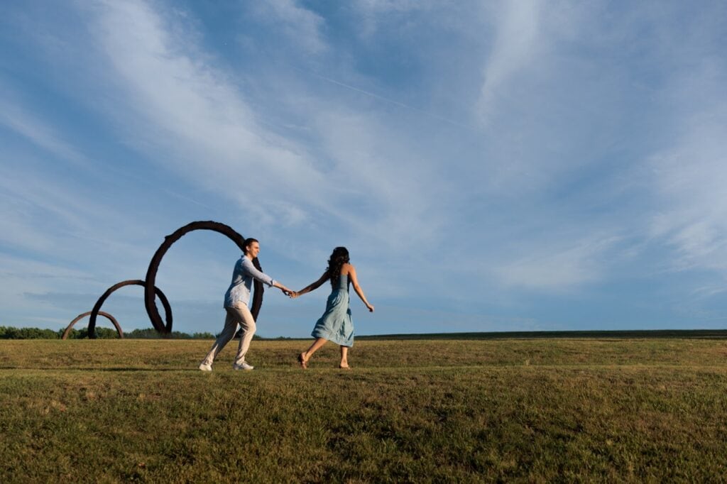 A couple holding hands while running in the filed near the Gyre sculptures at the NCMA in Raleigh. the sculpture is in the background as they have their photograph taken