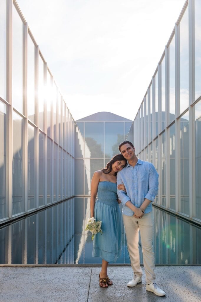 A husband and wife holding onto each other in front of the nc museum of art reflecting pool. They are modeled wearing matching blue and cream outfits while she hold a bouquet of white flowers for her anniversary.
