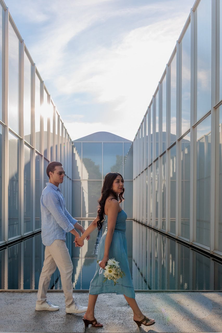 A couple walking in front of the reflecting pool at the NCMA in Raleigh. They are wearing blue matching outfits while she holds his hand and a bouquet of flowers