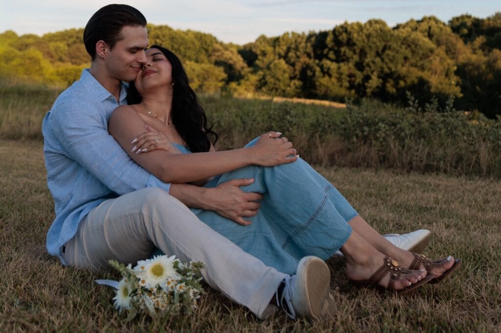 A couple sitting near tall grass a the NCMA in Raleigh NC. They have a bouquet of white flowers near their site while they hold each other during their anniversary photoshoot