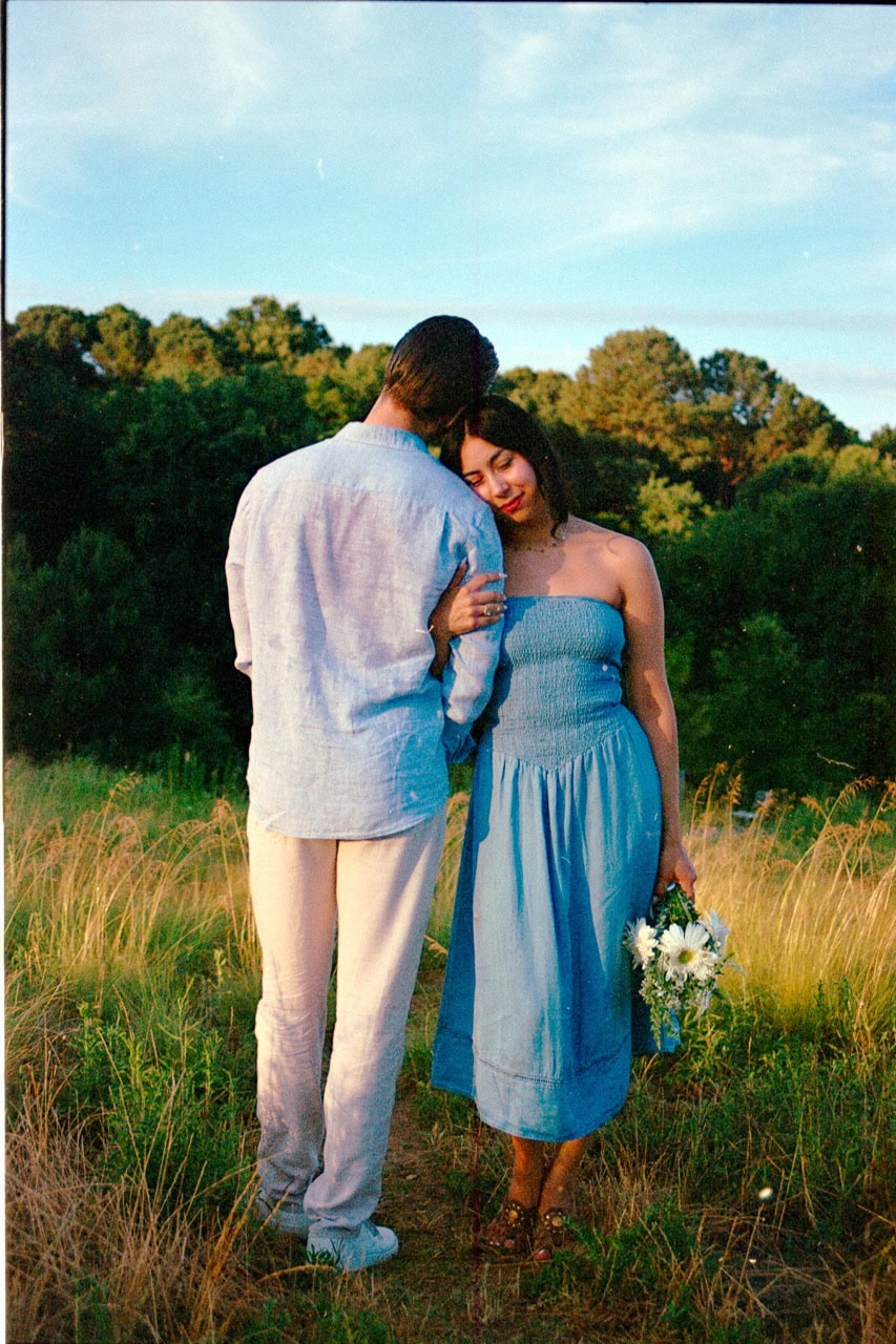 a 35mm film photograph taken of a couple standing in tall grass at the NC Museum of Art in Raleigh. The woman is looking down at the grass while holding a bouquet of flowers while her husband looks into the distance