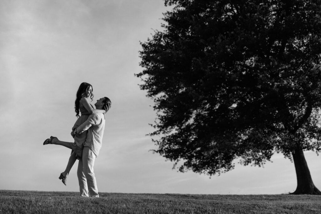 A black and white photograph of a couple standing in a field at the NC museum of art in Raleigh NC. They are standing near an oak tree in each others arms looking into each others eyes on their anniversary during a photoshoot with Amelia and Matt Photo