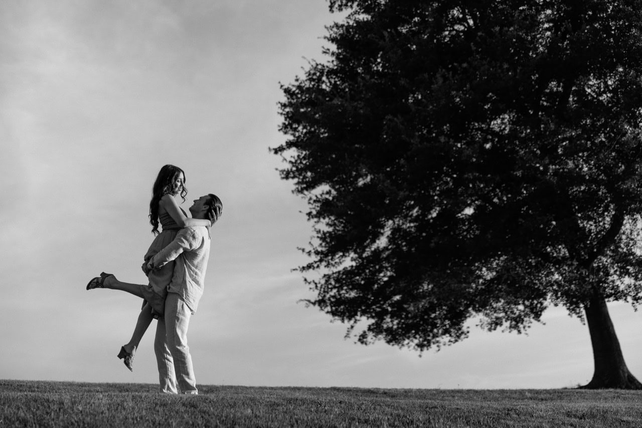 A black and white photograph of a couple standing in a field at the NC museum of art in Raleigh NC. They are standing near an oak tree in each others arms looking into each others eyes on their anniversary during a photoshoot with Amelia and Matt Photo