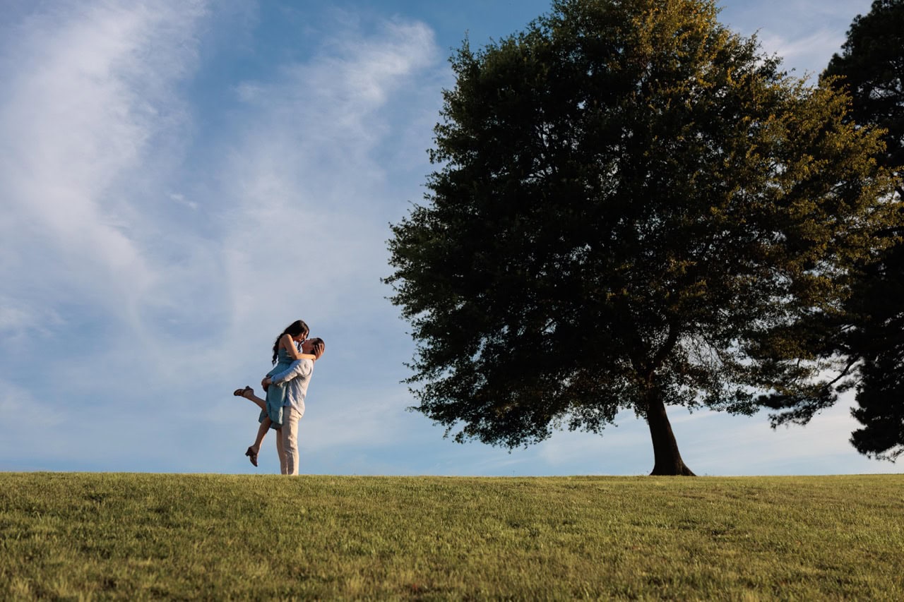 a man is lifting his wife during their anniversary photoshoot at the NC Museum of art. they are standing on a hill positioned under a large oak tree while standing in the grass kissing