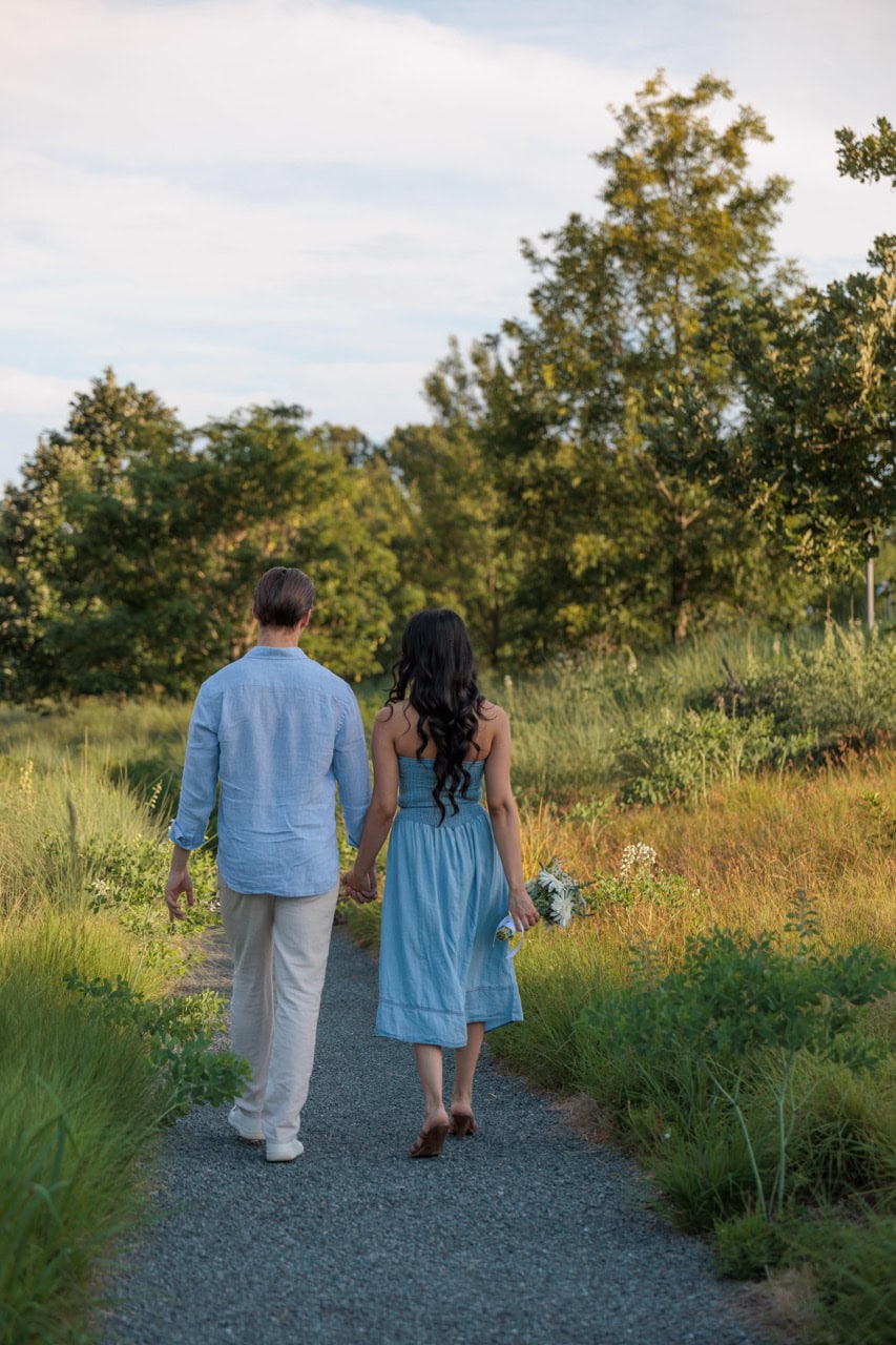 A couple walking amongst tall grass at the NC Museum of Art in Raleigh. They are walking to the field while wearing matching blue outfits. She is holding a bouquet of flowers.