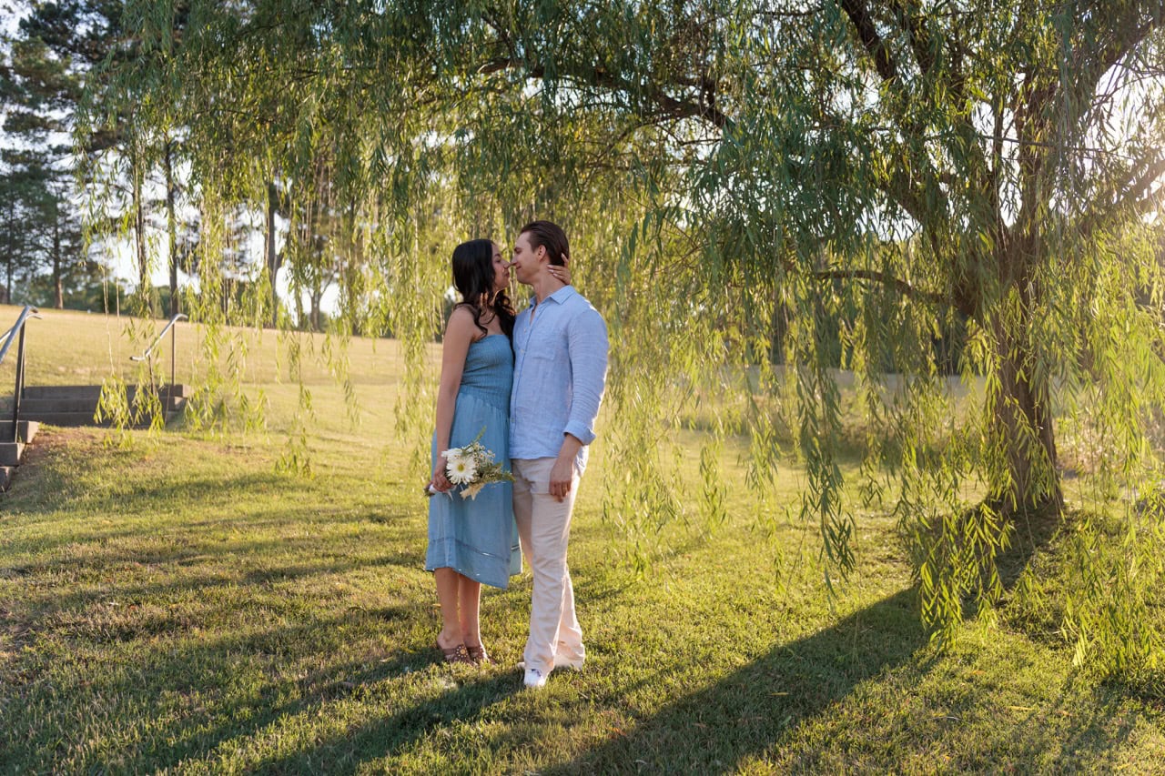 A couple standing in front of a weeping willow tree at the NC Museum of Art in Raleigh NC. They are posed looking into each others eyes while she holds his head. They are wearing matching outfits and she is holding a bouquet of flowers