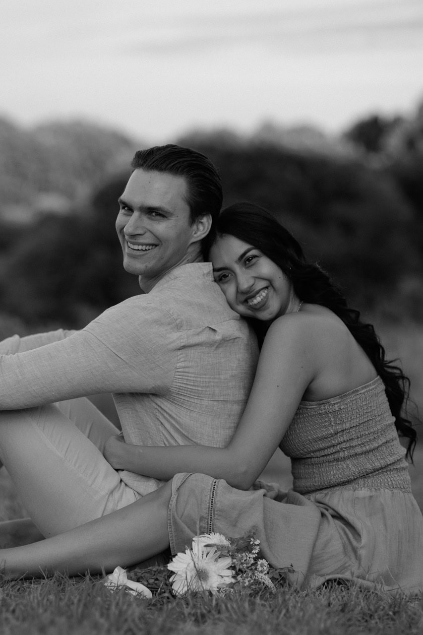 a black and white photograph of a couple sitting in the filed at the NCMA while they hold eachother and laugh