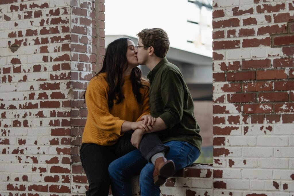 Couple sitting on a brick wall with fading bricks. They kiss and hold each other.