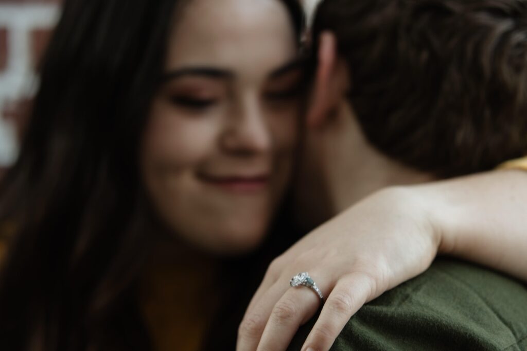 Couple sitting and hugging during their engagement session in Downtown Raleigh at Barcelona Wine Bar. She is showing off her engagement ring as he whispers into her ear.