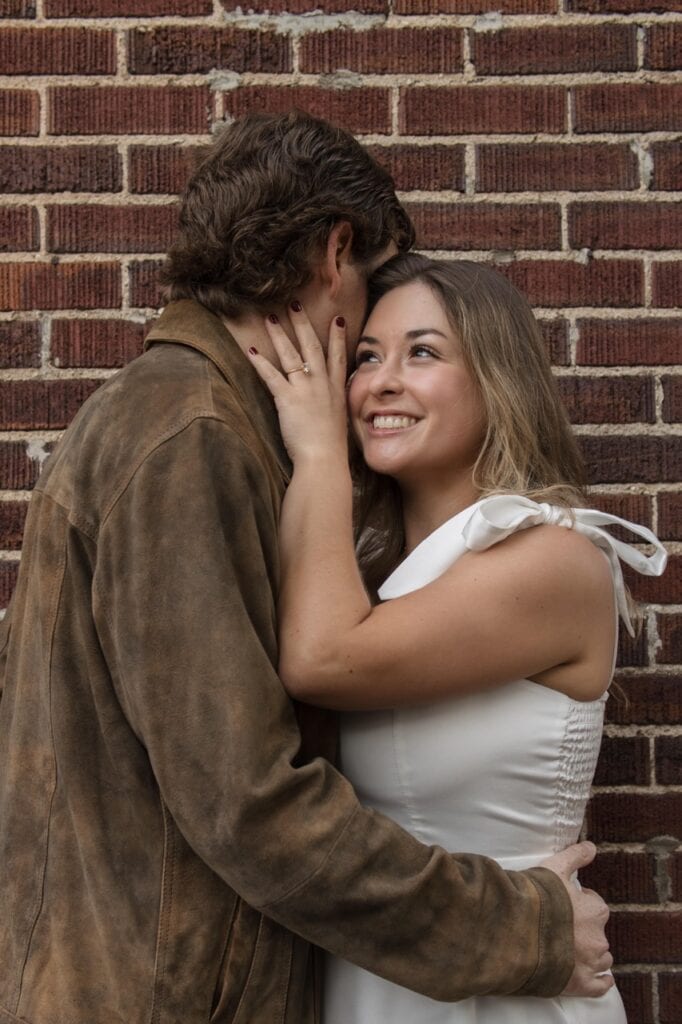 couple standing in front of a red brick wall in downtown raleigh in city market. She is wearing a white dress for the engagement session smiling as her fiancé whispers into her ear