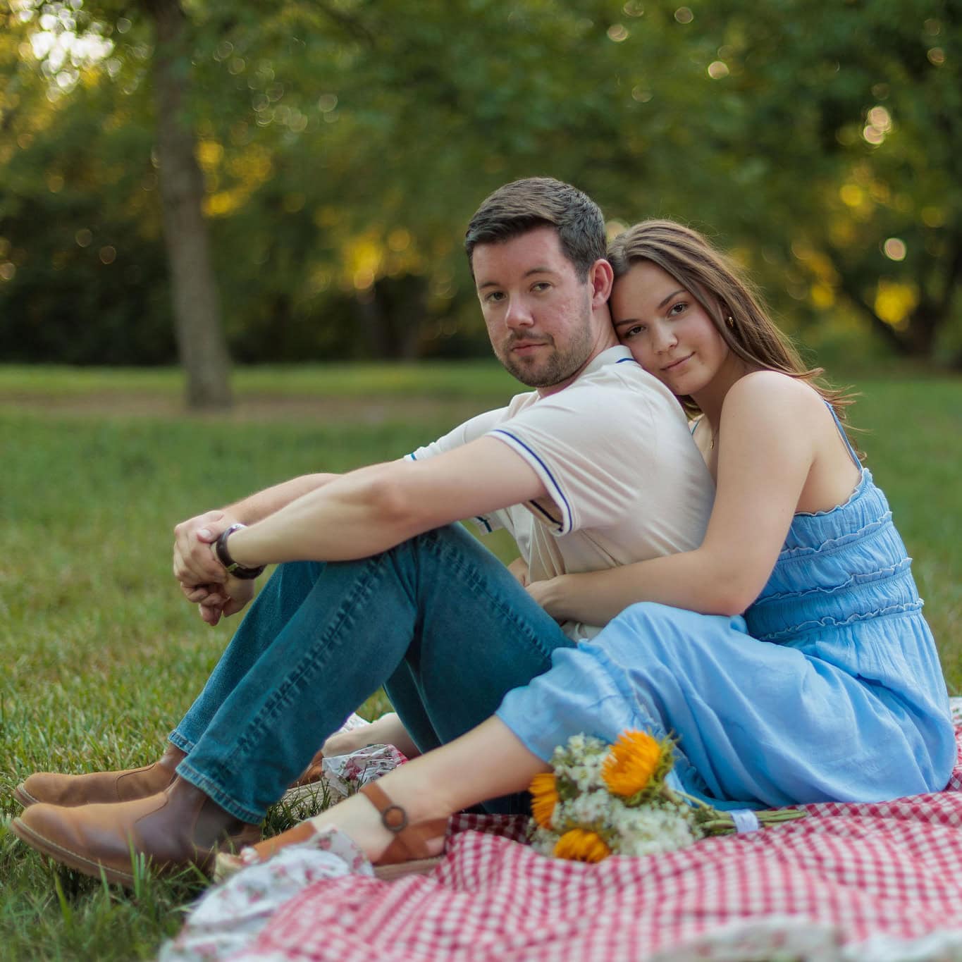 a couple looking into the camera sitting on a picnic blanket in a field at golden hour during their couples anniversary photography session with Amelia and Matt Photo at Historic Oak View Park in Raleigh NC