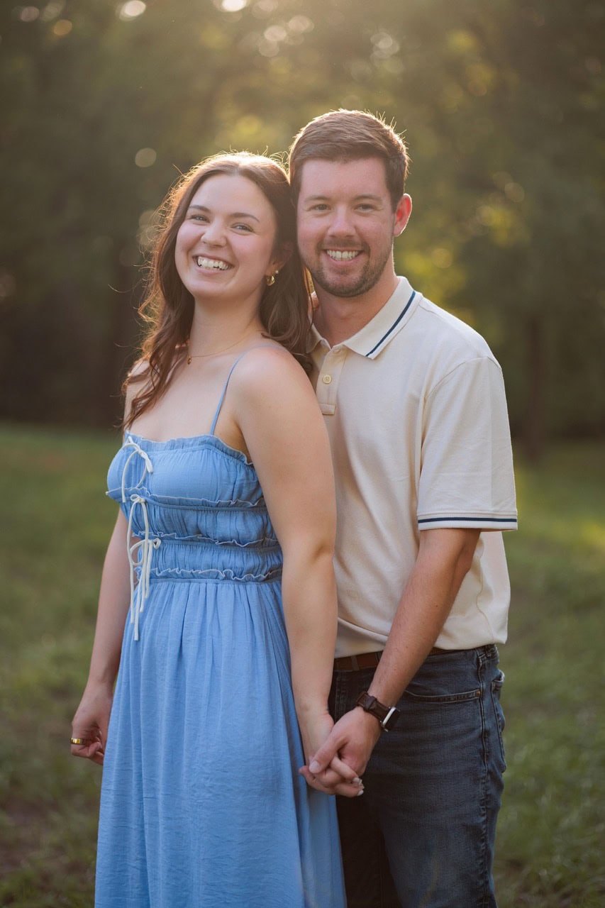 a husband and wife looking into the camera having their portrait taken at golden hour. She wears a blue dress while he has a white polo on. they are laughing and smiling