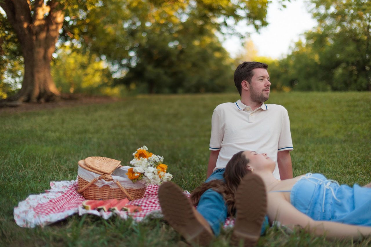 a wife resting her head on her husbands lets as they lay on a picnic blanket on a filed with trees behind them
