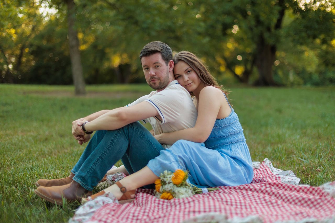 a wife holding her husband from behind as they sit on a picnic blanket with flowers next to them
