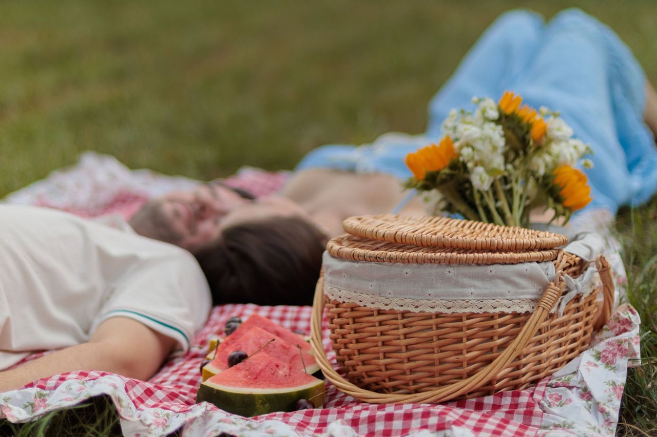 a couple laying on a picnic blanket with a picnic basket and watermelon in focus in front of them