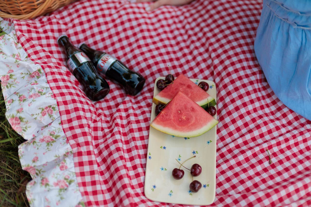 watermelon and cherries on a plate next to bottles of diet coke during a picnic on a blanket