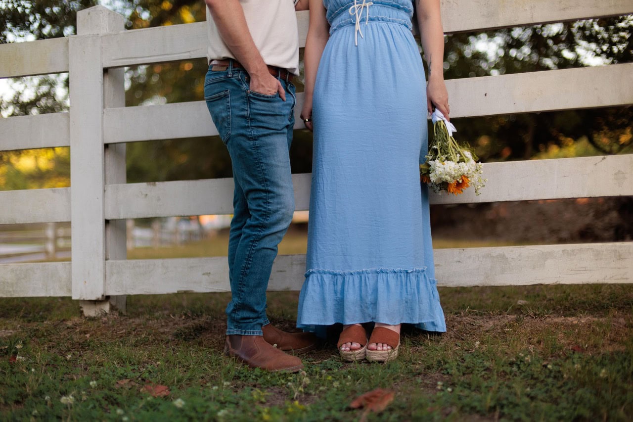 woman in a blue dress holding flowers standing next to her husband as they lean on a white fence