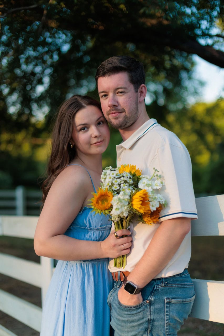 Couple looking into the camera posed for their couples session with Amelia and Matt Photo at Historic Oak View Park. They are leaning on a white picket fence leaning on each other holding flowers