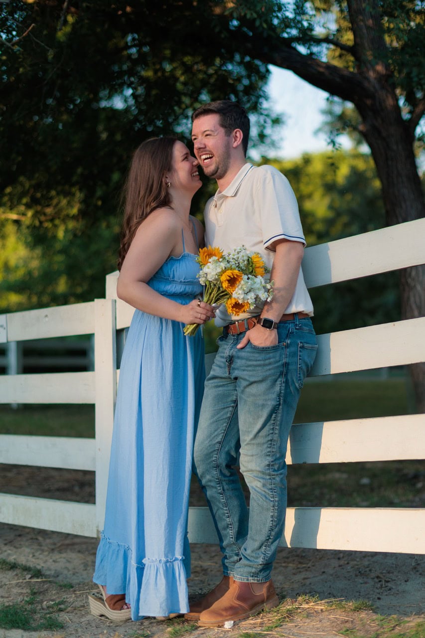 couple leaning on a white picket fence leaning into each other while holding flowers and looking into the camera at the couples photography session