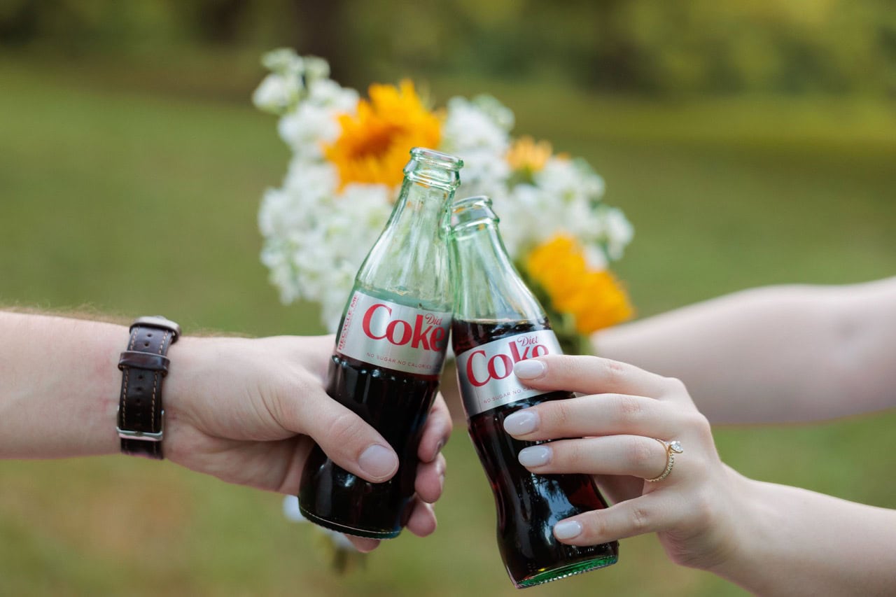 detail photograph of a couple touching the tips of diet coke bottles together while holding flowers