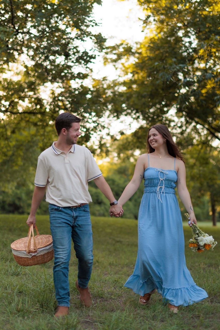 man holding a picnic basket while his wife holds flowers as they walk to their picnic at historic oak view park in raleigh nc at golden hour