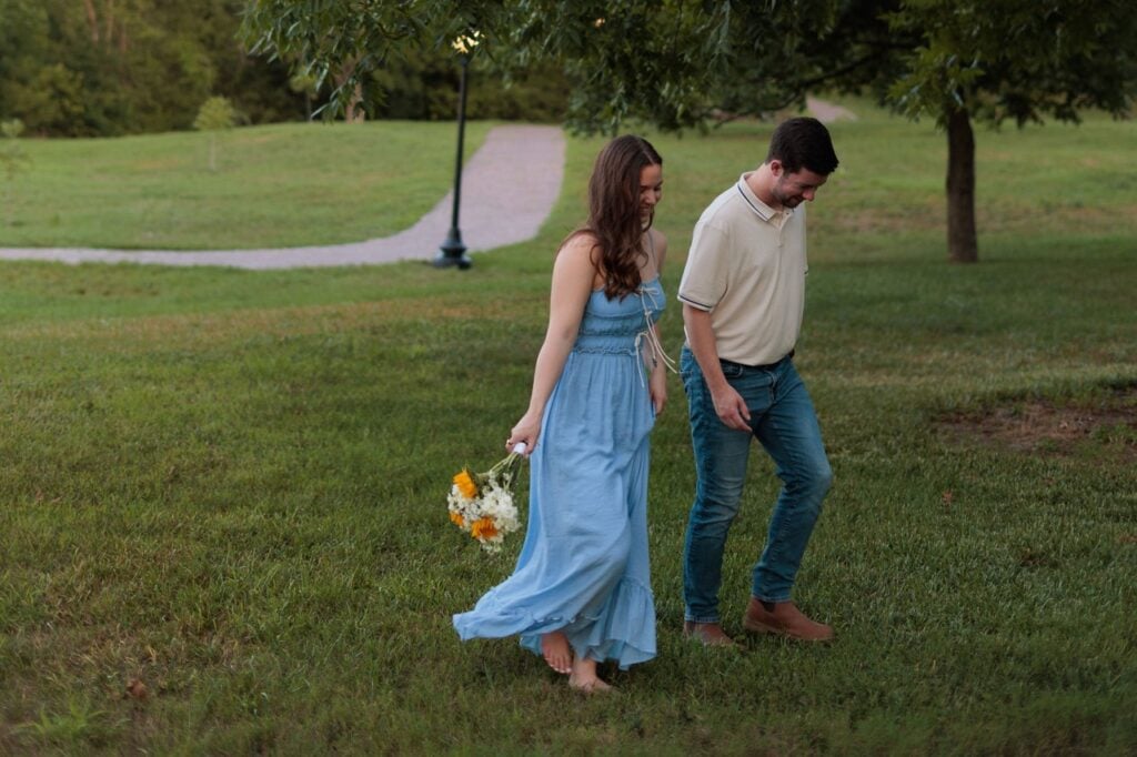 couple walking on the filed at the historic oak view park in Raleigh NC holding her husbands hand and some flowers while wearing a flowy blue dress