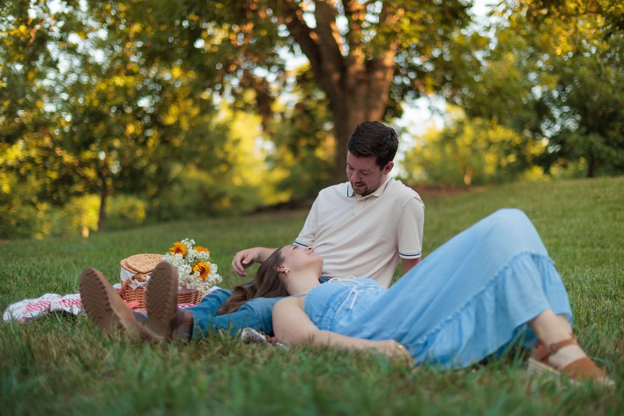 couple having a picnic, woman is leaning on her husbands lap and a picnic basket is next to them at golden hour