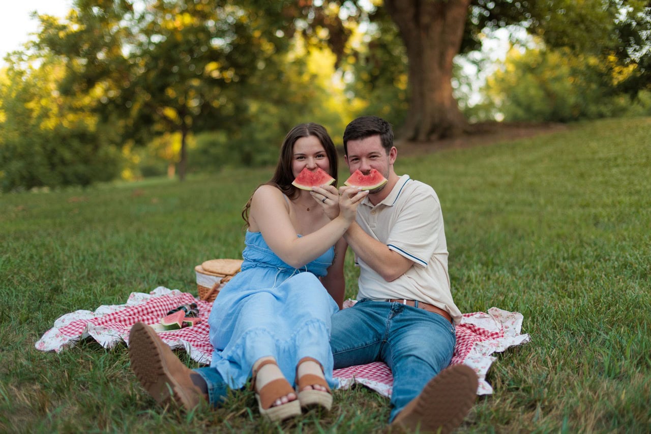 Couple sitting on a checkered picnic blanket in a field holding up watermelon in front of their faces during golden hour at a couples photography session at the Historic Oak View Park