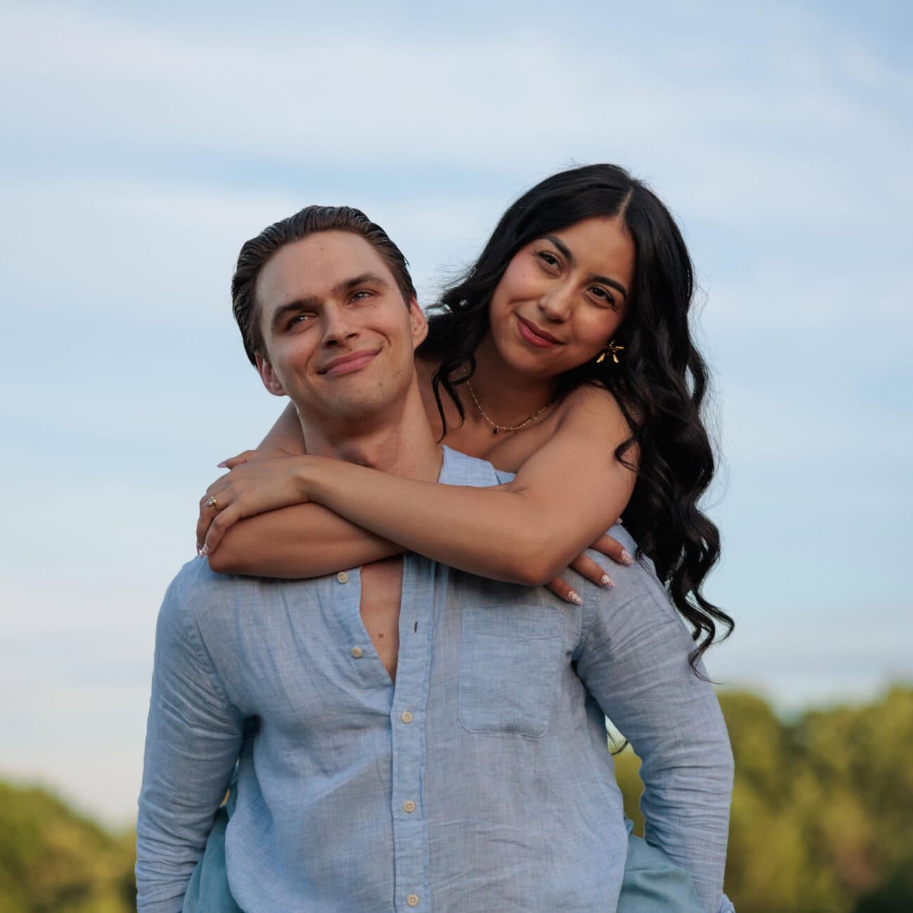 Man holding his wife on his back with the sky and clouds in the background at the NC Art Museum in Raleigh NC at Golden Hour For their Anniversary Photoshoot