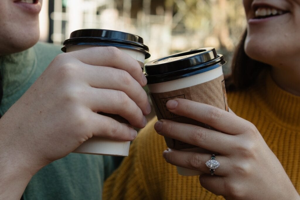 Couple tapping their coffees together showing off her enagement ring while smiling in downtown Raleigh