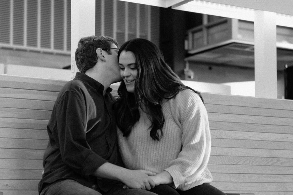 Couple sitting in the union station train station in downtown raleigh. It is a black and white photo where a man is whispering into his fiancé's ear