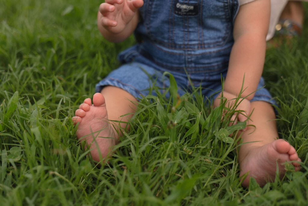 A baby sitting in the grass while wearing blue jean overalls and playing with the grass