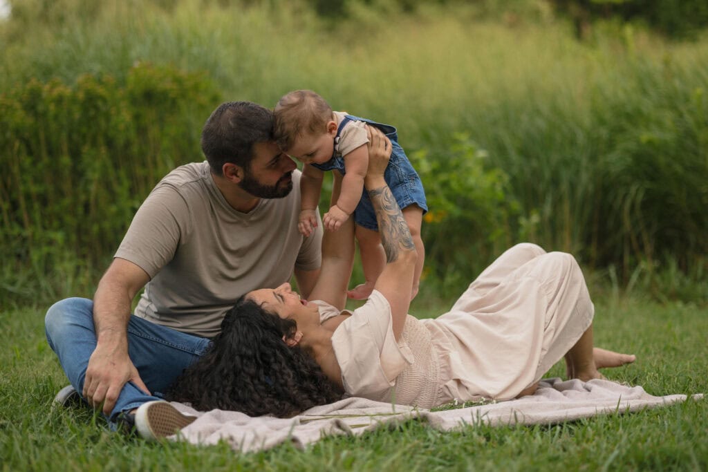 A mom laying on a picnic blanket in a field while holding up her baby as she and dad look at the baby