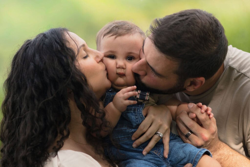 A 9 month old baby being kissed on the cheeks by his parents 