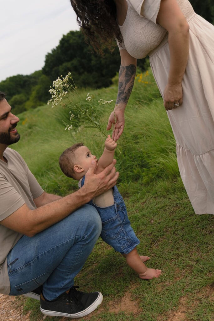 A 9 month old baby being held by his dad while handing flowers to his mom