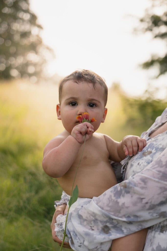 A baby smelling a flower while being held by his mother during their family photography session with Amelia and Matt Photo during golden out at the NC Museum of Art in Raleigh, NC